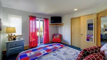 A bedroom with a bed, a television mounted on the wall, a desk with a lamp, and a window with blinds.