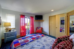 A bedroom with a bed, a television mounted on the wall, a desk with a lamp, and a window with blinds.