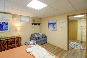 A room with a bed, a grey sofa, a wooden dresser, a wall-mounted shelf with plants, a painting, and a door leading to another room. - Sibley Shores Lake Cabin w/ Dock & Deck! (Pequot Lakes)