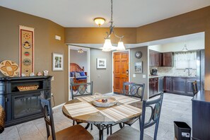 A dining area with a wooden table, chairs, and a chandelier. There is a kitchen visible in the background with dark cabinets and a window. - Desert-view Green Valley Retreat Near Golf Courses (Green Valley)