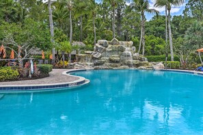 A pool with a rocky waterfall feature surrounded by palm trees and a brick walkway. - Olé at Lely Resort, More Than a Vacation (Naples)