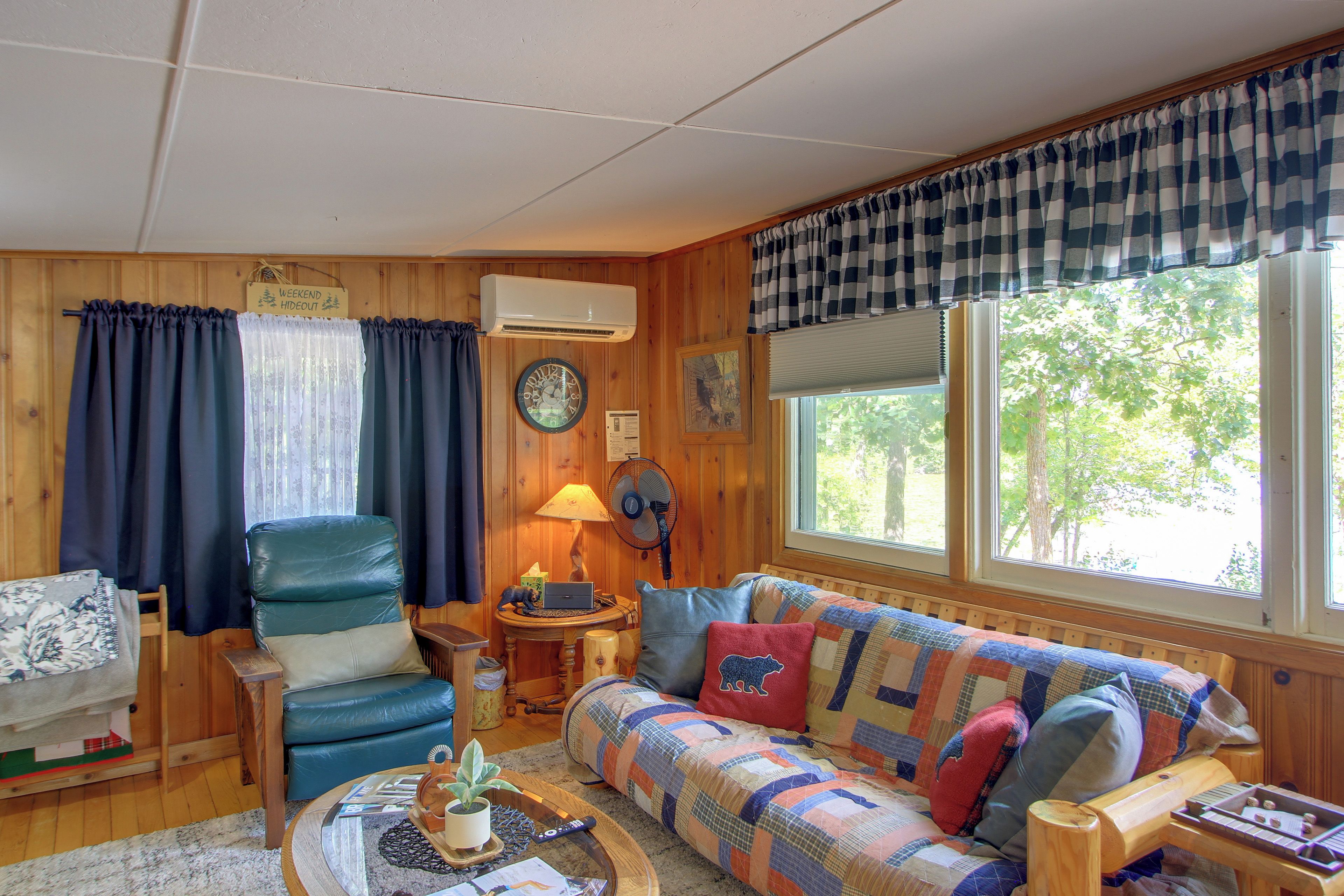 A cozy cabin interior with a wooden interior, a sofa, a chair, a table with a lamp, and a clock on the wall.