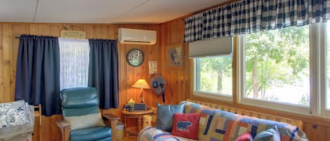 A cozy cabin interior with a wooden interior, a sofa, a chair, a table with a lamp, and a clock on the wall.
