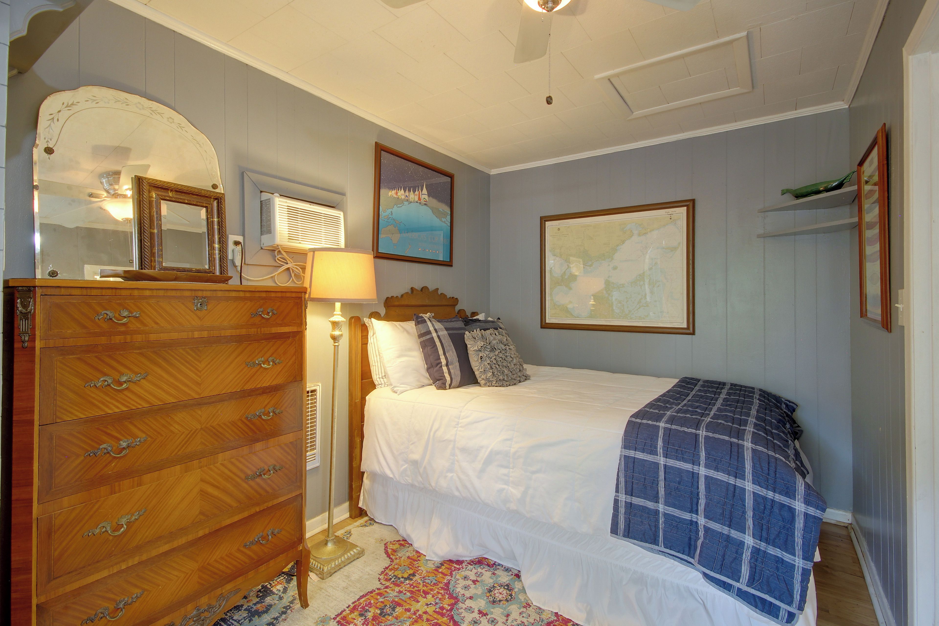 A bedroom with a wooden dresser, a bed with a blue blanket, a map on the wall, and a lamp.