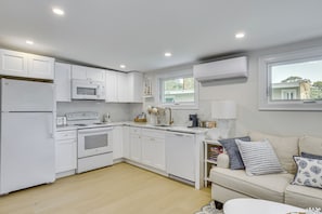 A modern kitchen with white cabinetry, a built-in microwave, a stove, and a dishwasher. There is a window above the sink, and a white sofa with cushions in the adjacent living area. - Sun, Surf & Seafood: York Home 2 Blocks to Beach (York Beach)