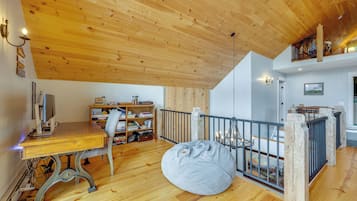 A room with a wooden ceiling, a desk with a computer, a bookshelf, a bean bag, and a view of a mountain through a window.