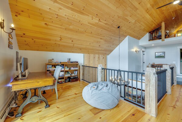 Una habitación con techo de madera, un escritorio con computadora, una estantería, un sillón de peluche y una vista de la montaña a través de una ventana.
