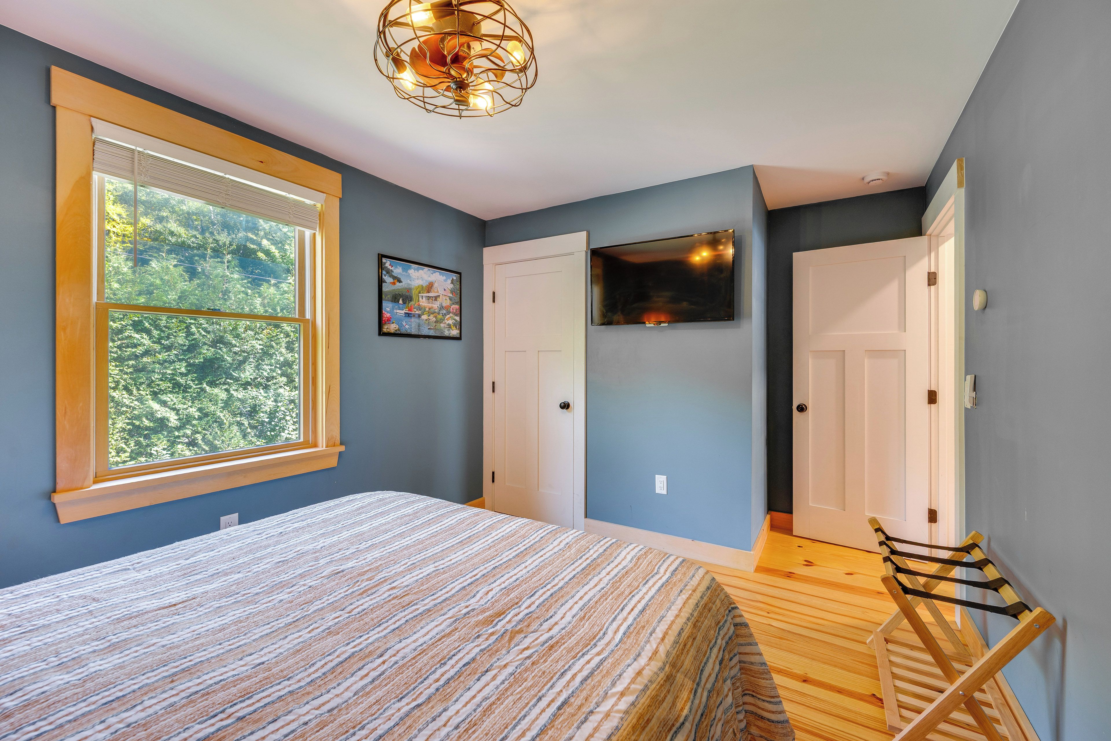 Un dormitorio con cama, ventana con persianas, un cuadro, televisor y puerta.