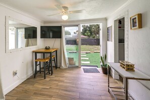 A room with a ceiling fan, a wooden table with two chairs, a television mounted on the wall, and a view of a backyard with a wooden fence and greenery. - 6 Mi to Beach! Sarasota Studio w/ Screened Porch (Sarasota)