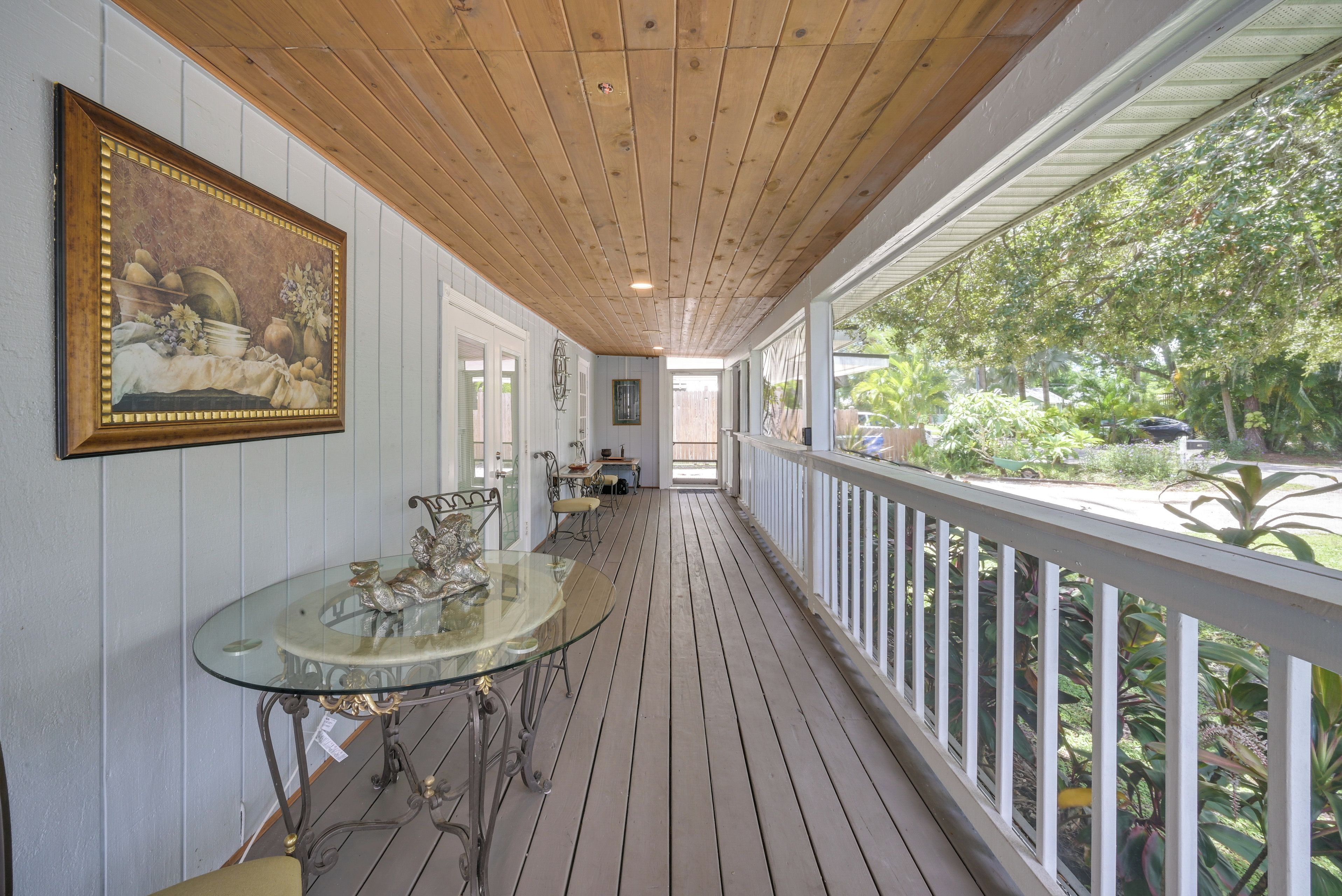 A covered porch with a glass table and chairs, a painting on the wall, and a view of greenery outside.