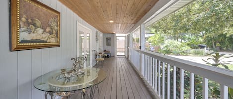 A covered porch with a glass table and chairs, a painting on the wall, and a view of greenery outside.