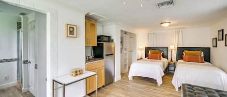 Habitación de hotel compacta con kitchenette, dos camas, una mesa pequeña y un ventilador de techo.