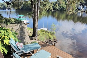 A wooden deck with two lounge chairs and a small table, overlooking a calm lake with a dock and a birdhouse.