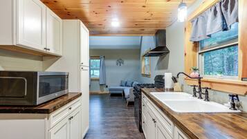 Una cocina compacta con armarios blancos, microondas, lavamanos con grifo antiguo y ventana con vistas a un bosque.