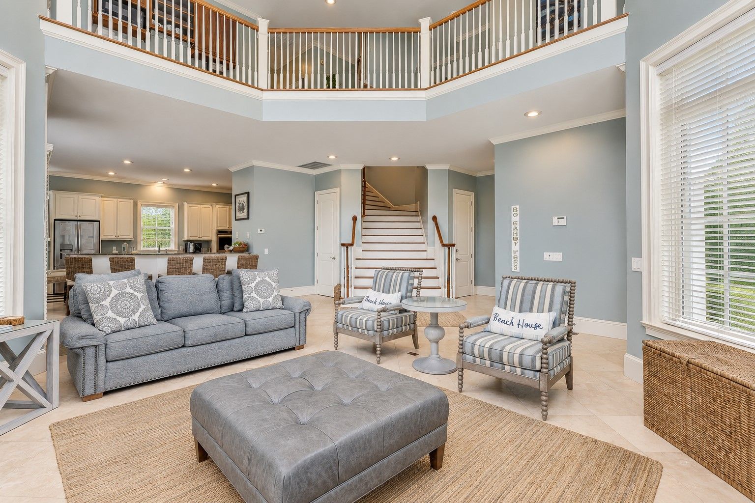 Un espacio habitable amplio con un sofá gris, sillones a rayas y un taburete gris. Una escalera con barandillas de madera conduce a un nivel superior.