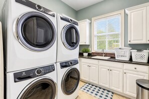 A laundry room with stacked front-loading washers and dryers, a window with blinds, a sink, and a potted plant. - 20 Knotts Way: North Forest Beach Retreat w/ Private Pool, Spa & Beach Access (Hilton Head Island)