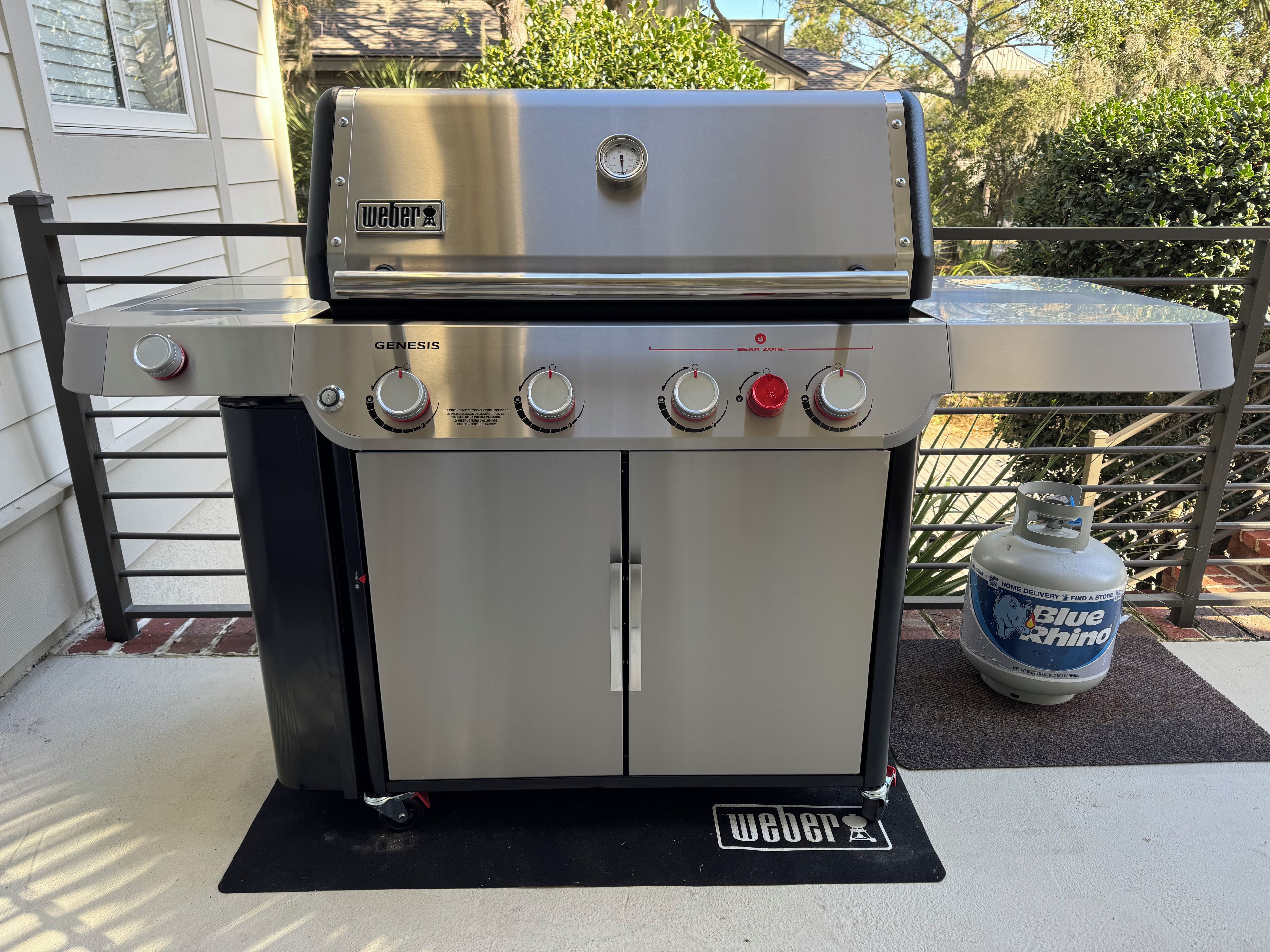A stainless steel Weber Genesis gas grill with four control knobs, a built-in thermometer, and a gas tank nearby.