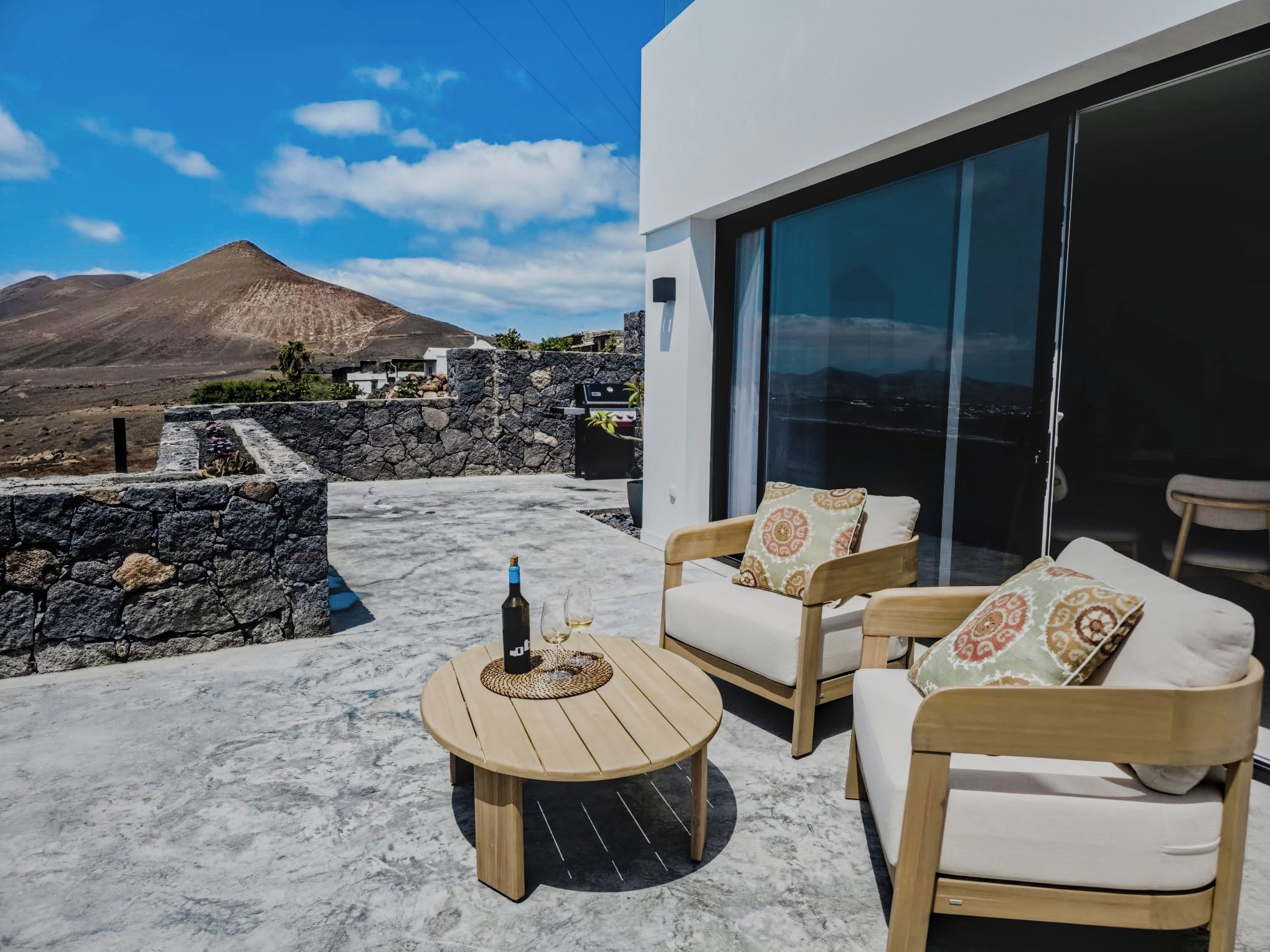 A modern outdoor seating area with a stone wall, a wooden table with a bottle and glasses, and a view of a mountain.