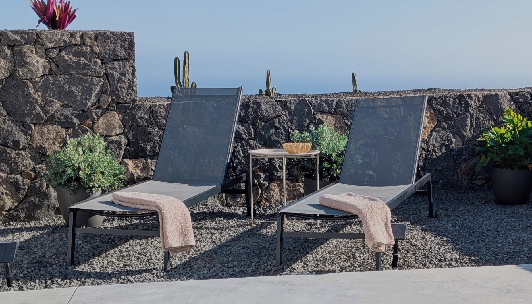 Two lounge chairs with towels, a small table with a bowl, and a potted plant, set against a stone wall with cacti.