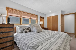 Een slaapkamer met een groot bed, een houten hoofdbord, een raam met zonwering, een nachtkastje met een lamp en een kledingkast.