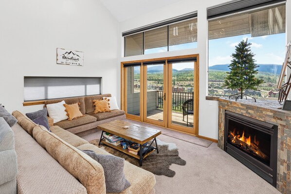 A modern living room with a sectional sofa, a coffee table, a fireplace, and large windows offering a scenic view.