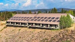 Ein Gebäude mit mehreren Solaranlagen auf dem Dach, umgeben von einer trockenen Landschaft mit vereinzelter Vegetation und fernen Bergen.
