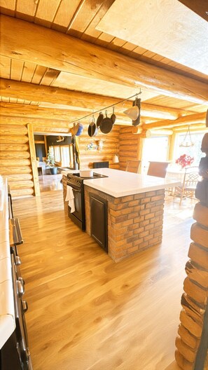 L’intérieur d’une cabane en rondins présente un espace cuisine avec un îlot central en briques, un plancher en bois et des ustensiles de cuisine suspendus.