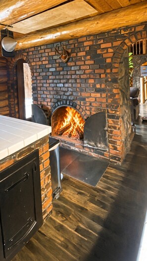 A brick fireplace with a fire, a wooden ceiling, and a tiled floor. - The Chief's Cabin Tumalo 4BR Log Cabin with Cascade Views (Bend)