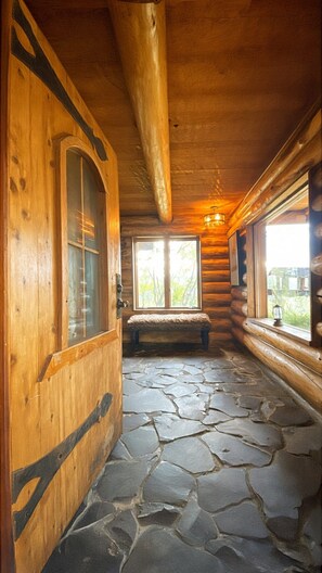 A log cabin interior with a stone floor, wooden walls, a bench, and a window with a view.