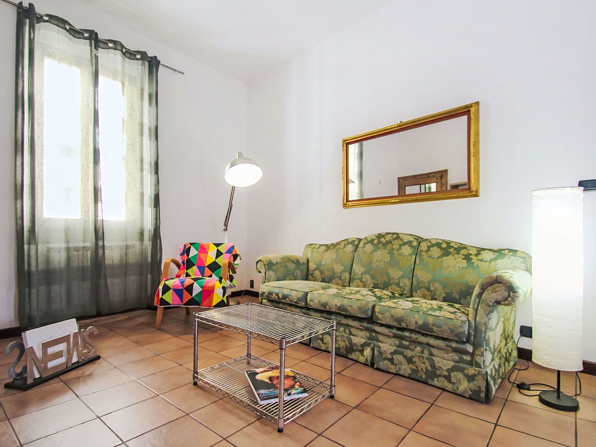 A living room with a sofa, a colorful chair, a metal coffee table, a lamp, and a mirror on the wall.