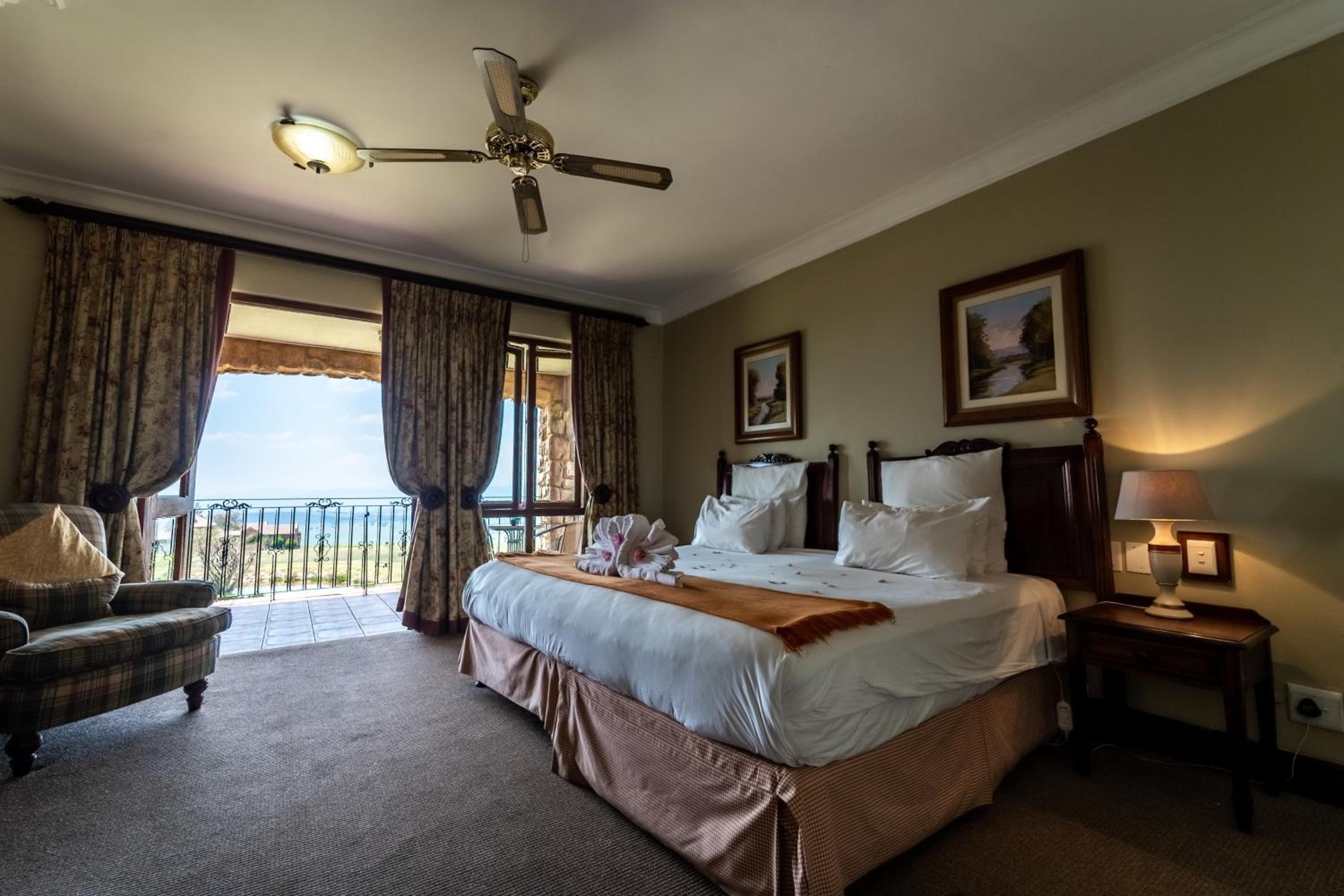 A hotel room with a large bed, a ceiling fan, a view of the sea, a chair, and a nightstand with a lamp.