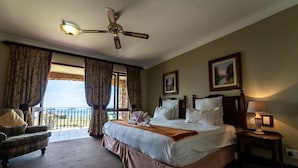 A hotel room with a large bed, a ceiling fan, a view of the sea, a chair, and a nightstand with a lamp.