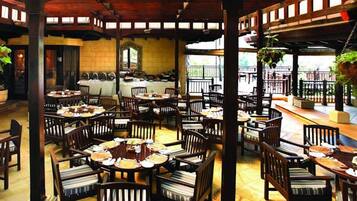 An indoor dining area with multiple tables and chairs, a kitchen in the background, and a wooden ceiling structure.