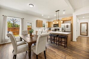A modern kitchen with wooden cabinets, a central island with bar stools, and a dining area with a large table and chairs. - Lake House Game Garage - Fire Pit Sleeps 8 (Granbury)