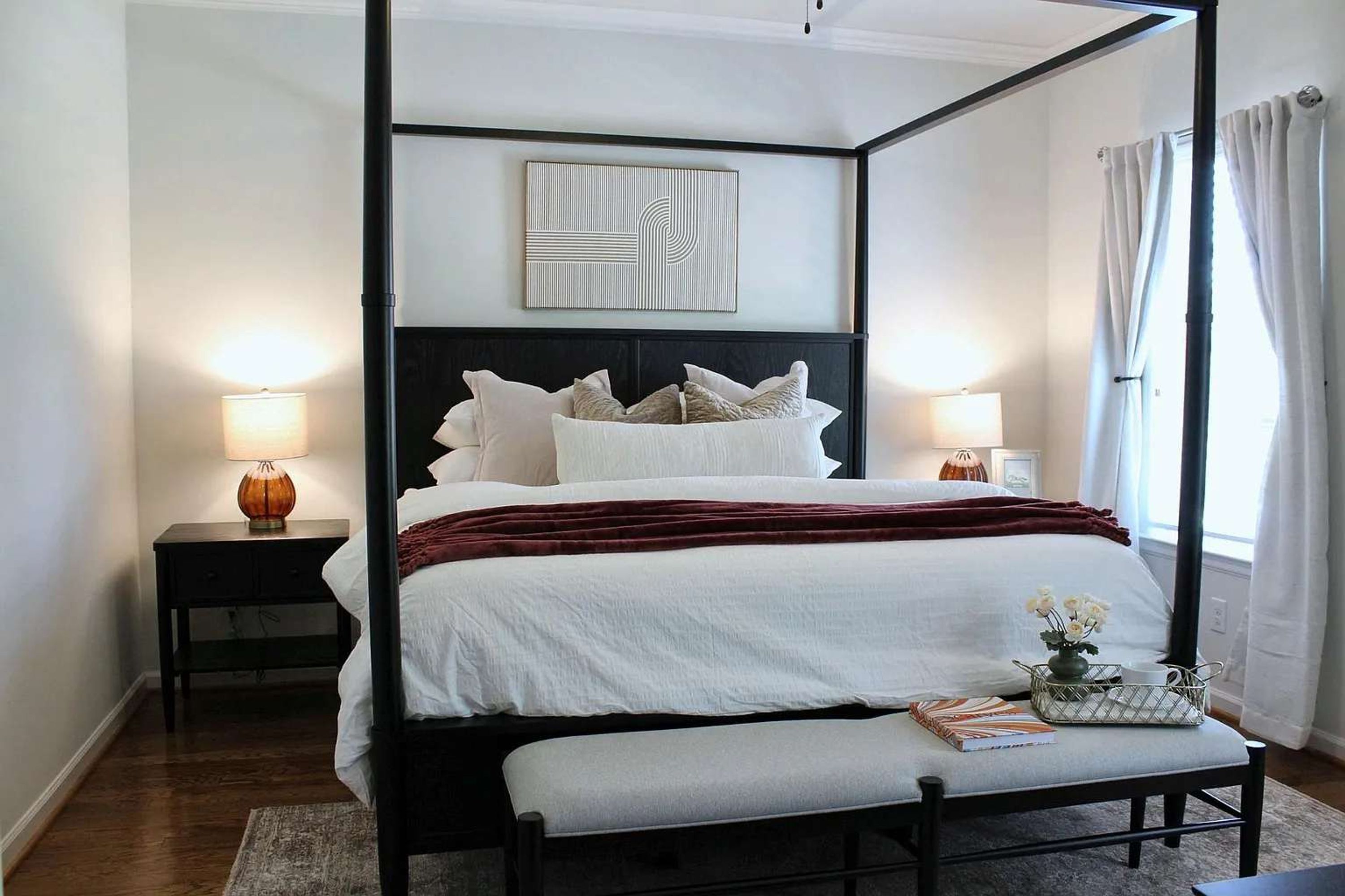 A four-poster bed with a dark frame, white bedding, and multiple pillows. There is a bedside table with a lamp on each side of the bed. A bench is placed in front of the bed.