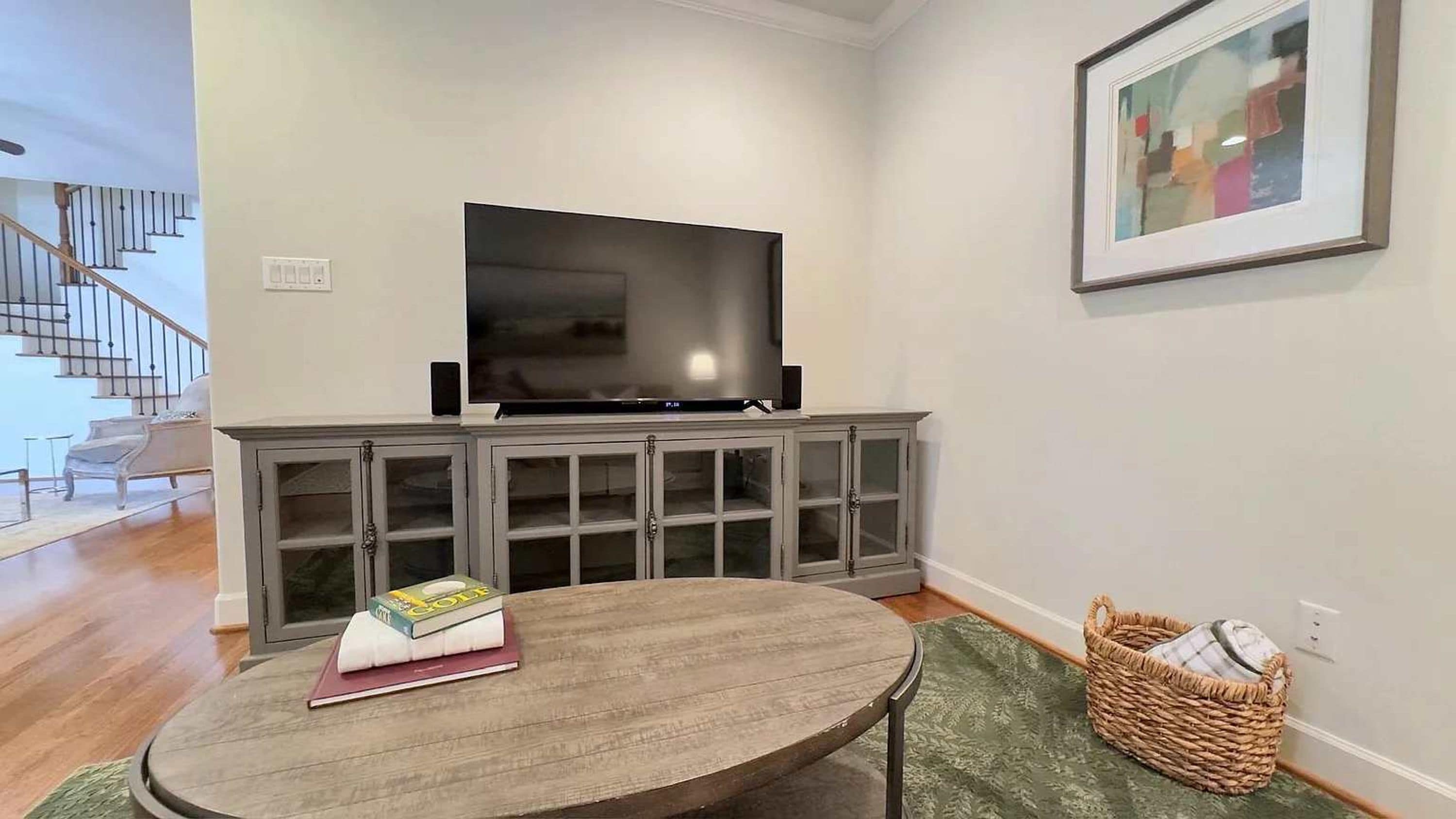 A living room with a flat-screen TV on a cabinet, a round table with a book, and a wicker basket.