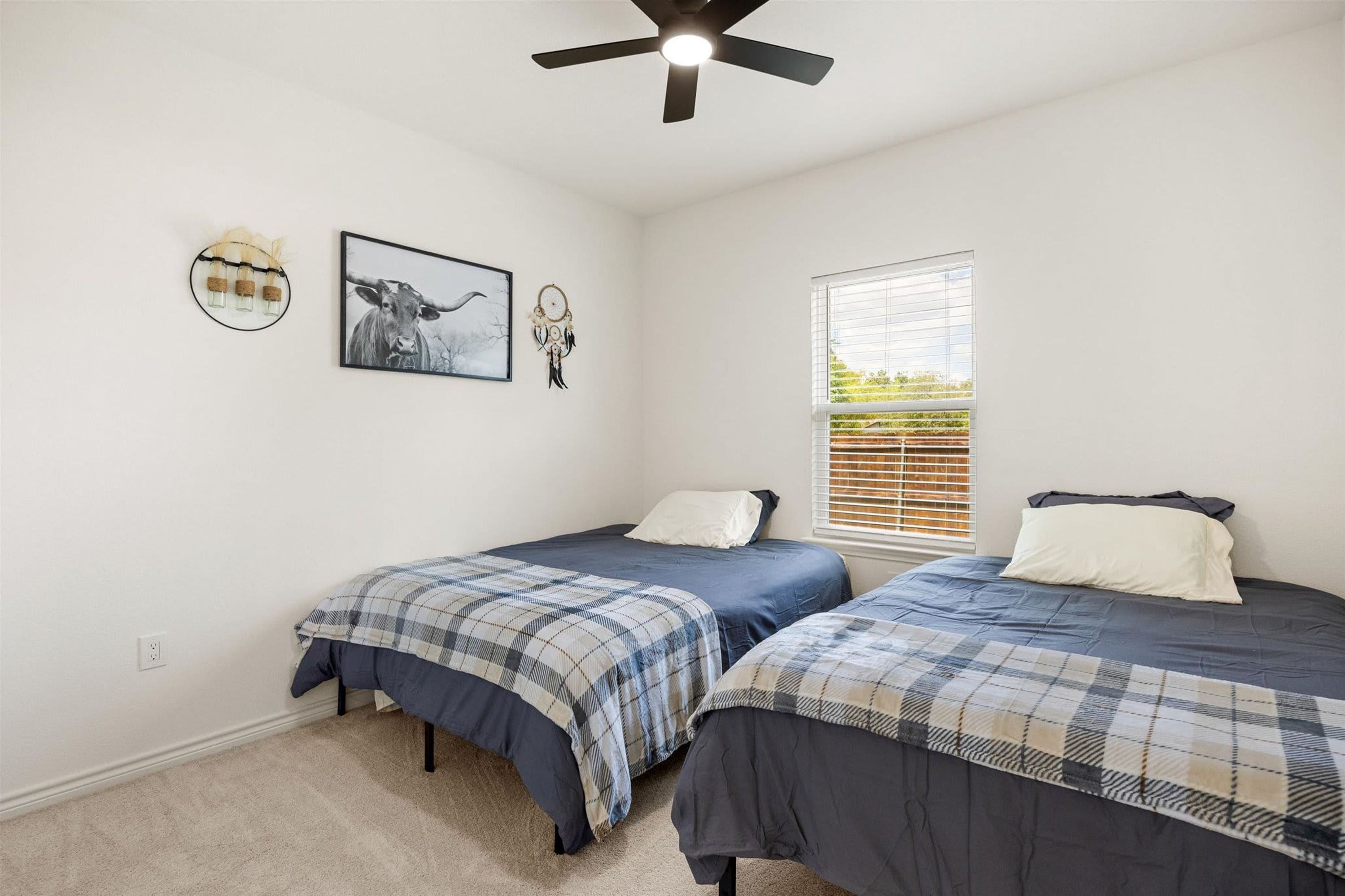 A bedroom with two beds, a ceiling fan, a window with blinds, and wall decorations including a framed picture and a dreamcatcher.