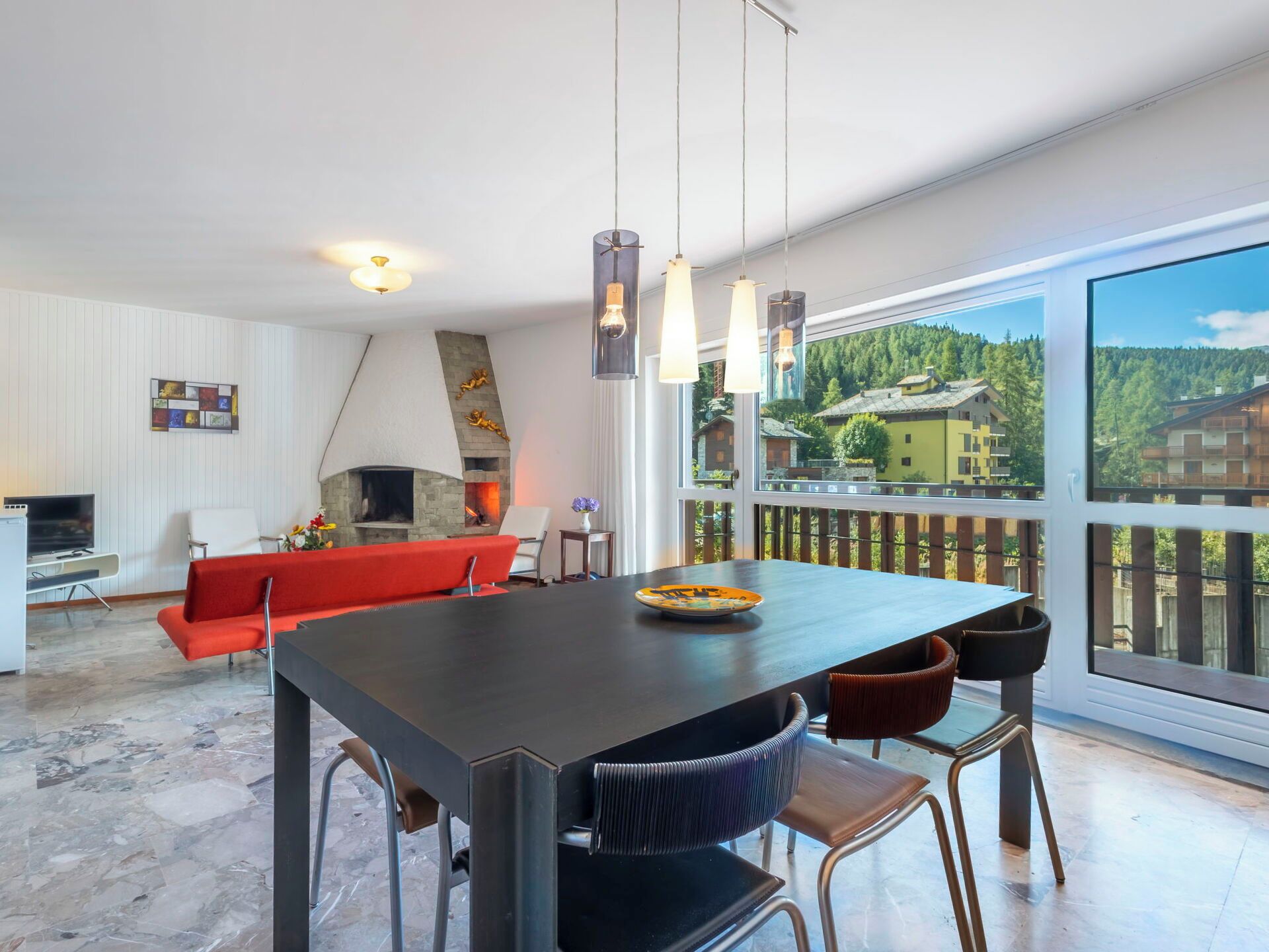 Un espacio habitable moderno con comedor, sofá rojo, chimenea y vista a un paisaje montañoso.