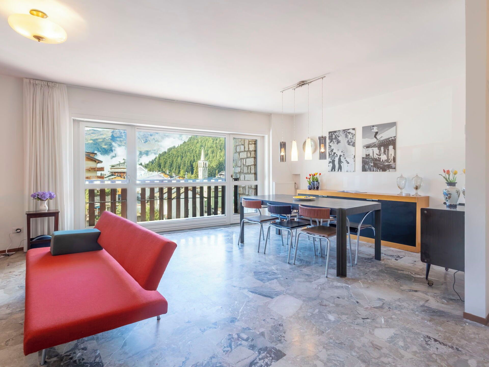 Una sala de estar moderna con un sofá rojo, un comedor con mesa y sillas, una encimera de cocina y un balcón con vistas a las montañas.
