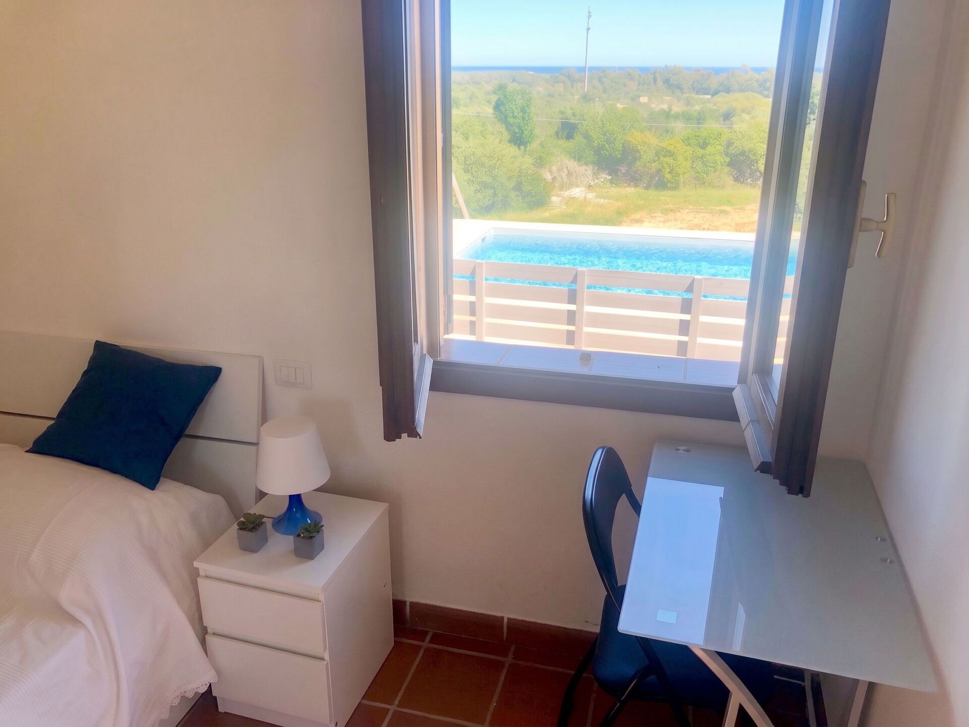 A bedroom with a bed, a desk, a chair, a nightstand, a lamp, and a view of the sea through a window.