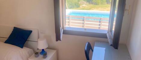 A bedroom with a bed, a desk, a chair, a nightstand, a lamp, and a view of the sea through a window.