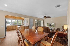 A spacious dining area with a wooden table and chairs, a ceiling fan, and a view of the kitchen through large windows. - Walk to Lake & Village | Hot Tub, Fire Pit, Fenced Yard & Lake Views (Big Bear Lake)