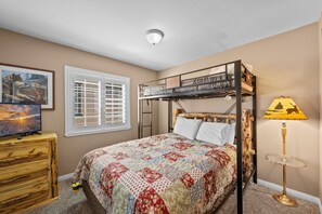 A bedroom with a bunk bed, a desk with a computer, a lamp, a window with blinds, and a painting on the wall. - Big Bear Bunkhouse | Hot Tub, Game Room, A/C, EV Charging & Fenced Yard (Big Bear City)