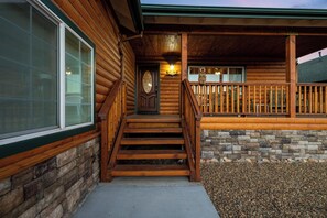 A wooden cabin with a stone foundation, a front porch with a railing, and a staircase leading to the entrance. - Big Bear Bunkhouse | Hot Tub, Game Room, A/C, EV Charging & Fenced Yard (Big Bear City)