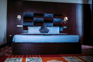 Una habitación de hotel con una cama grande, cabecera de madera oscura, dos lámparas de pared y una alfombra estampada.
