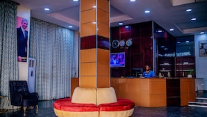 Eine Lobby mit einer zentral gelegenen Sitzgruppe in Rot und Beige, einem Empfangstresen und einem Fernsehbildschirm.