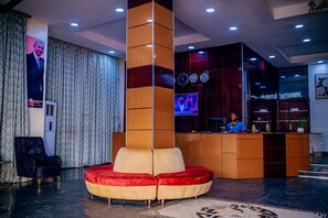 A lobby with a central red and beige seating area, a reception desk, and a television screen. - Dejavu Hotel and Suite (Akure)