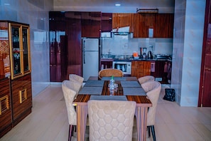 A dining area with a wooden table and cushioned chairs, a kitchen with wooden cabinets, and a built-in refrigerator. - Dejavu Hotel and Suite (Akure)