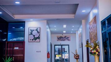 Un hall de hotel con mostrador de recepción, una escalera que sube a los pisos superiores y una entrada con puerta de vidrio.