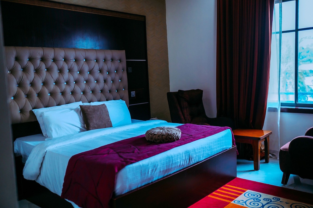 A bedroom with a tufted headboard, a bed with white and blue bedding, a purple throw blanket, and a small wooden side table.